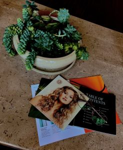 Magazines and photographs spread out on a table with a pair of reading glasses reflecting the photographer.